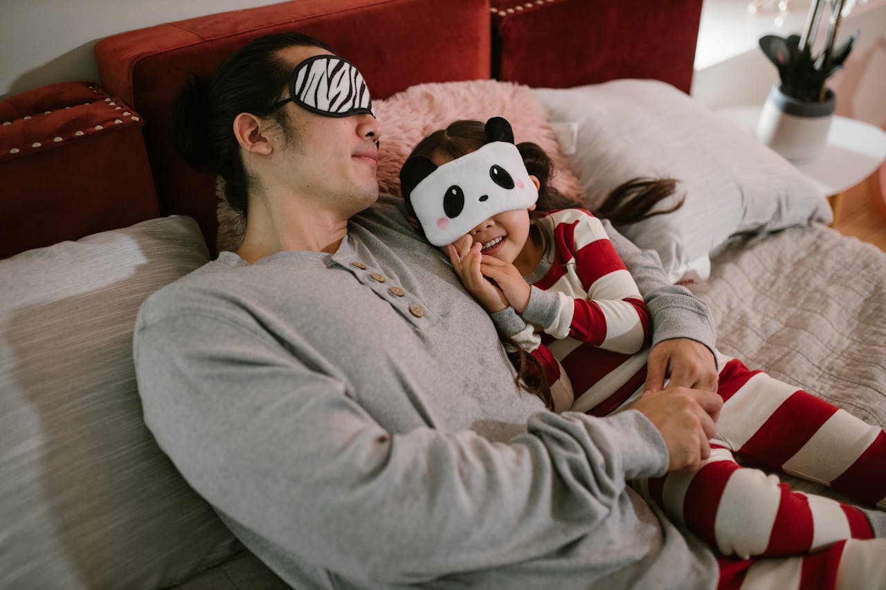 A father and daughter wearing cute sleep masks, sharing a tender bedtime moment indoors.