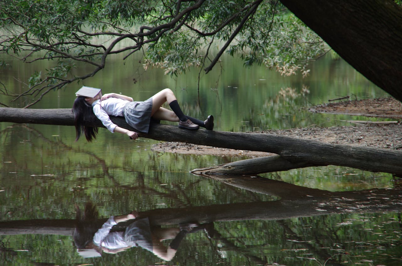 A young woman with a book on her face lies relaxing on a tree trunk by a tranquil lake.