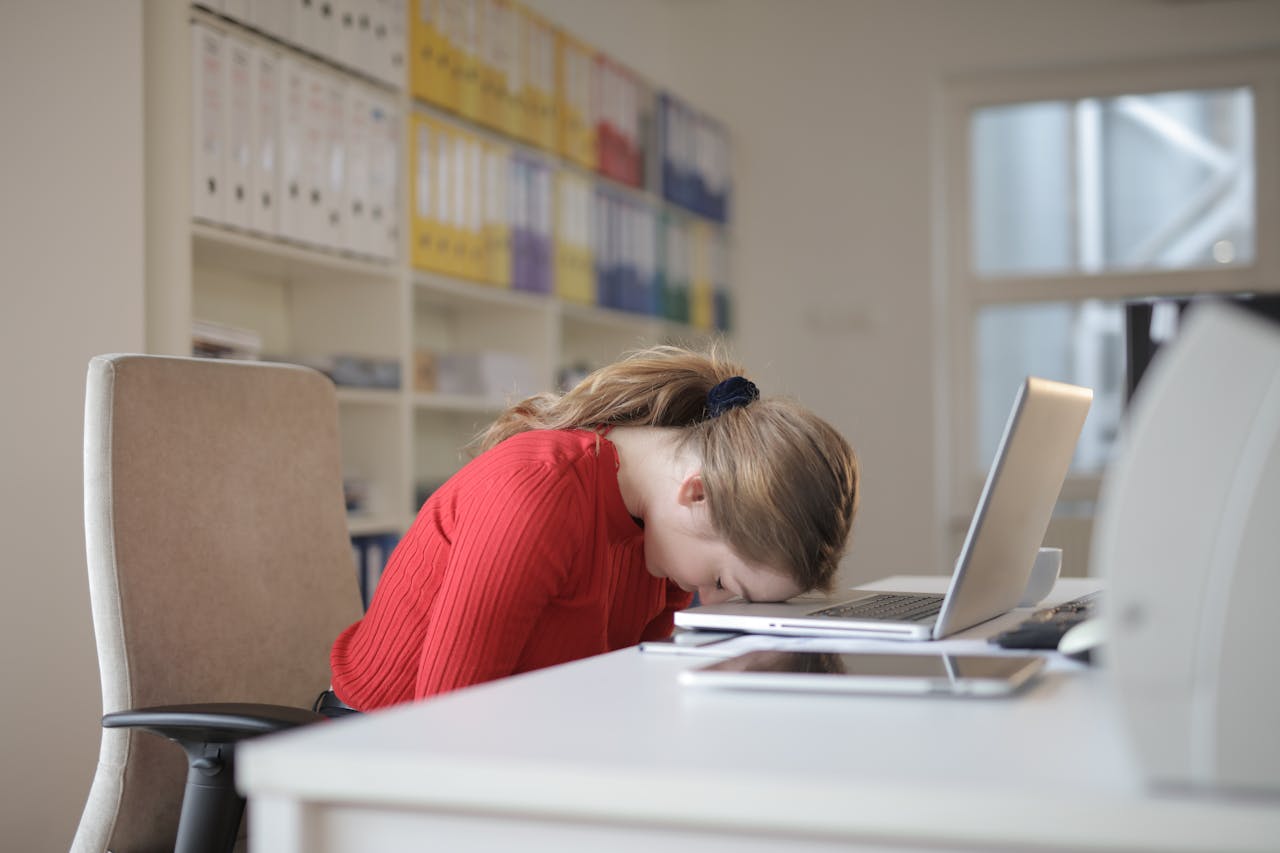 A tired woman in a red sweater leans her head on a desk with a laptop, symbolizing workplace fatigue.