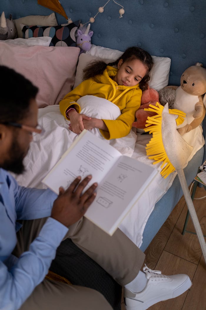 A father reading a bedtime story to his daughter tucked in bed with her stuffed toys.